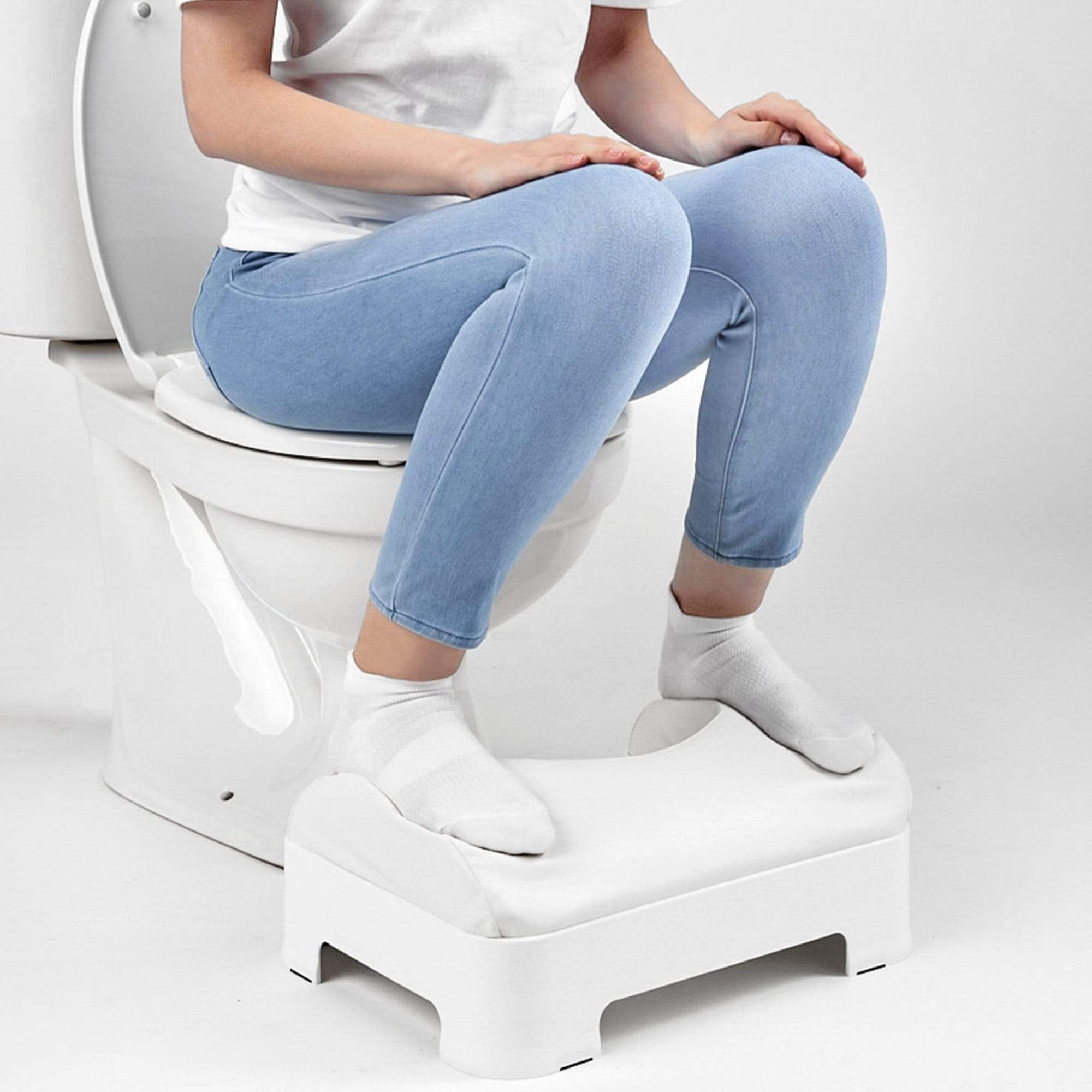 Model in a white shirt and jeans is sitting on the toilet while planting feet on the LUXE footstool