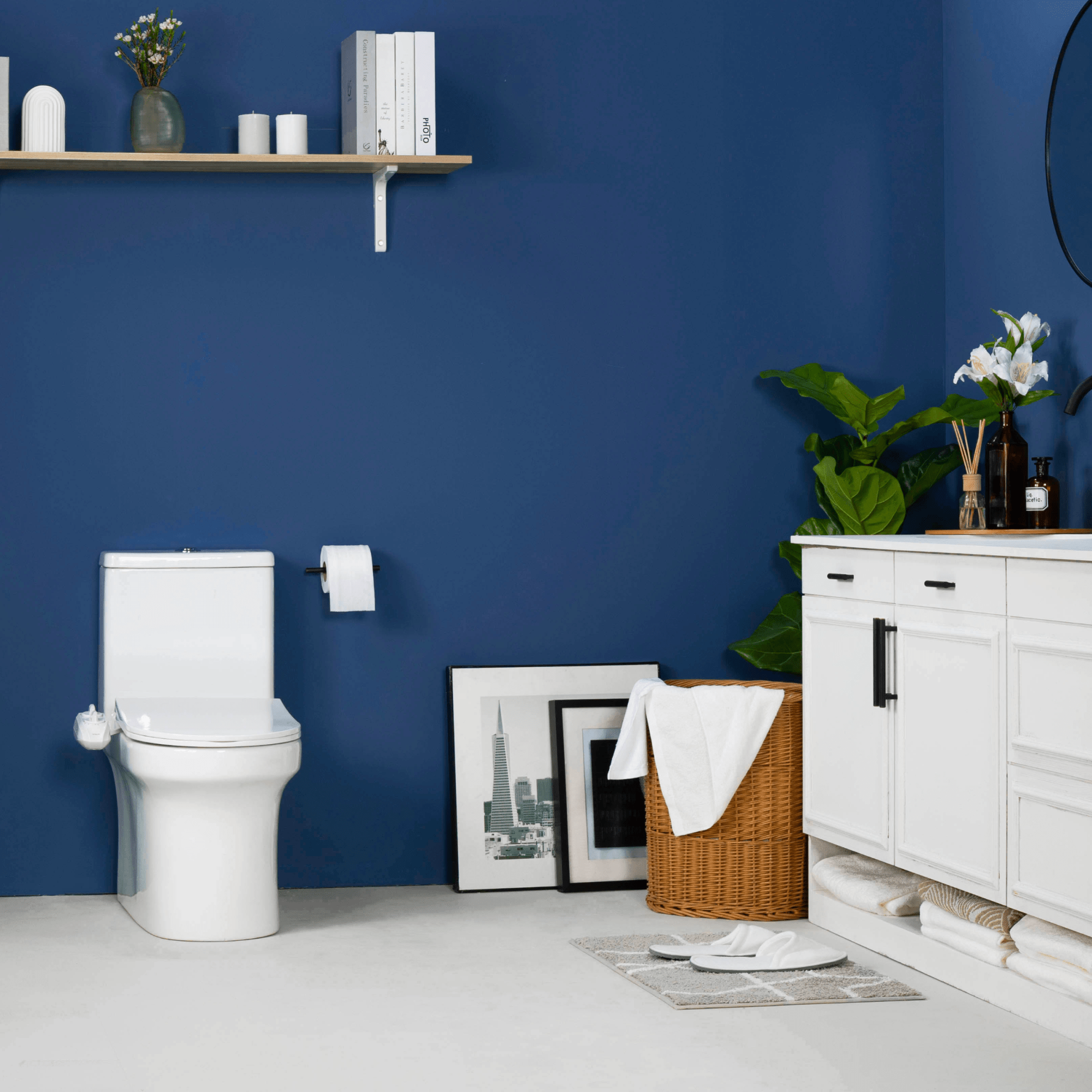 NEO 185 Plus White installed on a toilet, in a modern blue bathroom with a cabinet, paintings, and basket of towels